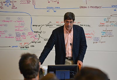 A man stands at a podium. A whiteboard behind him is covered with a complex diagram in dry erase marker