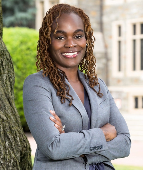 Ms. Breland-Noble is pictured wearing a gray suit and standing with her arms crossed with a tree, greenery, and building in the background.