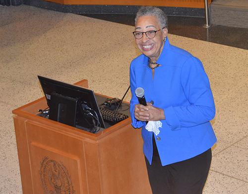 Dean Joy Williams stands beside a podium at the front of an auditorium, grasping a microphone in one hand