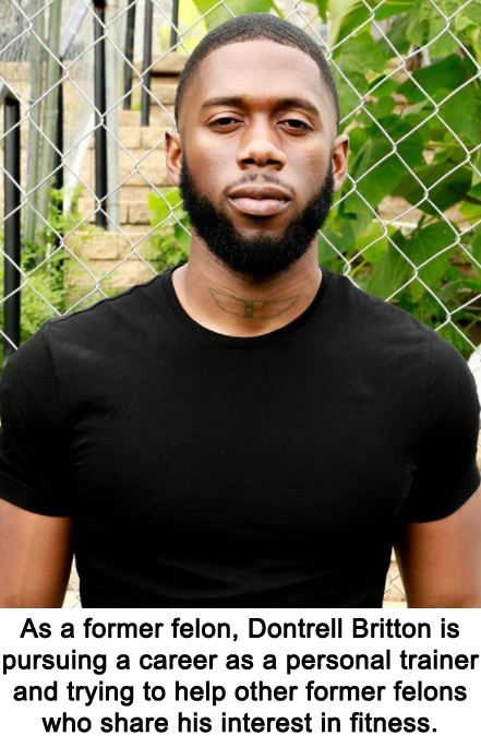 In this portrait shot a bearded black man stands against a chainlink fence behind which are green plants and a wall