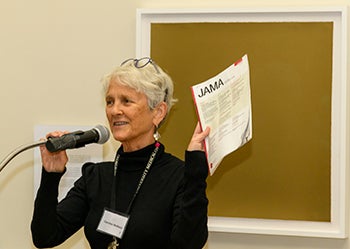 Caroline Wellbery holds up a copy of her journal article as she speaks into a microphone at a podium