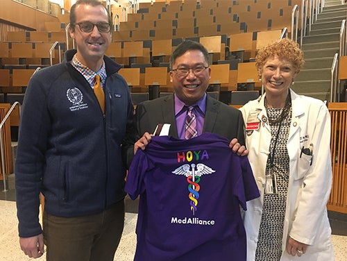 Three individuals stand next to each other in an auditorium setting, the center individual holds a purple Hoya MedAlliance shirt