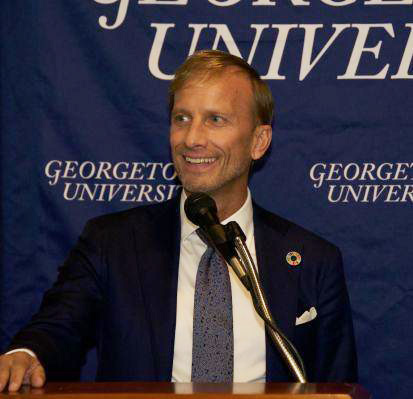 Mary Dybul smiling in front of a microphone in front of a blue banner that says "Georgetown University"