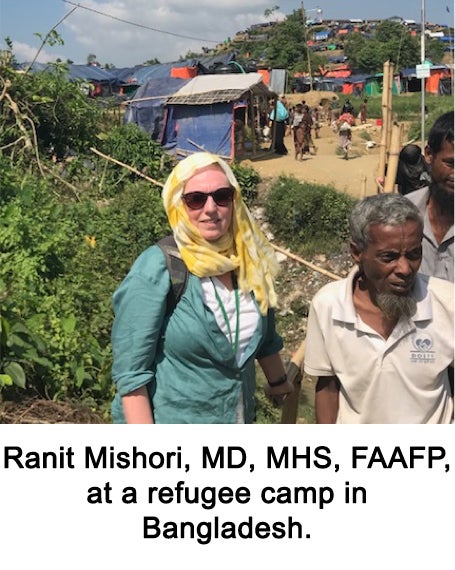 A woman wearing a headscarf stands next to a man in a refugee camp with a tent in the background