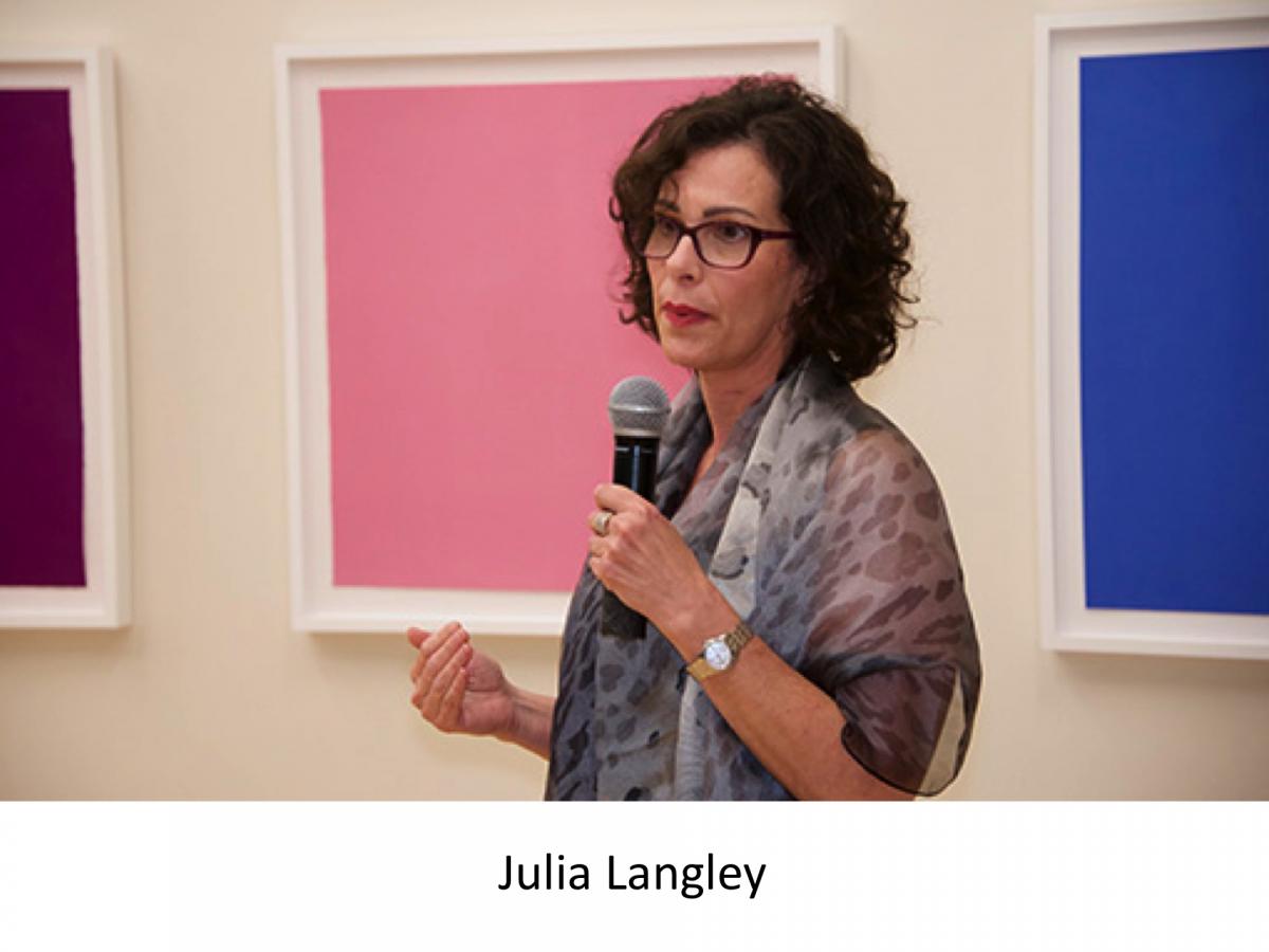 Julia Langley talking into a microphone in front of a sold baby pink print hanging on the wall