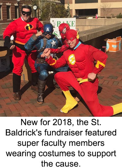 Four individuals pose wearing superhero costumes at an outdoor campus location