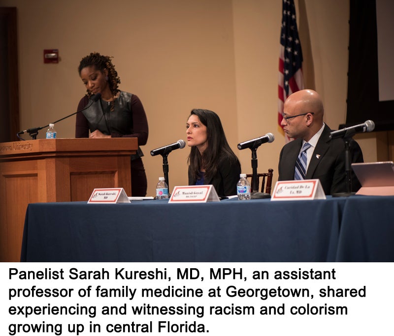 A man and woman sit behind a table. A woman stands behind a podium near them. The seated woman speaks into a microphone.