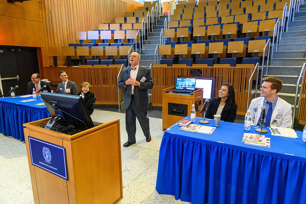 Several people sit at tables or stand in an auditorium