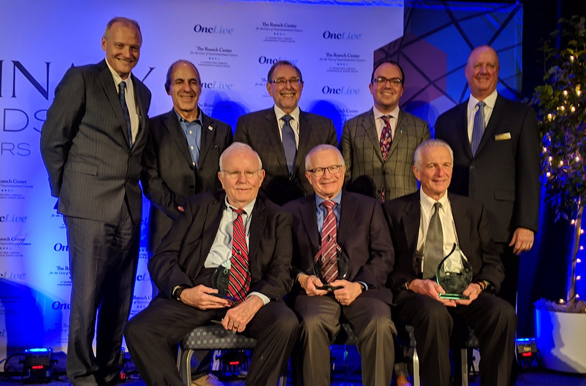 A group of men smiles onstage; three men in the front row hold crystal awards.