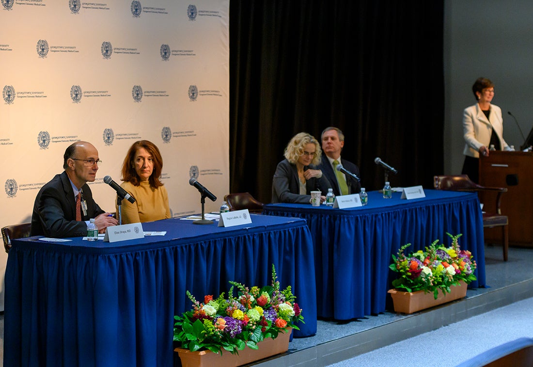 People sit at a long table draped in a blue tablecloth; a woman stands at a podium