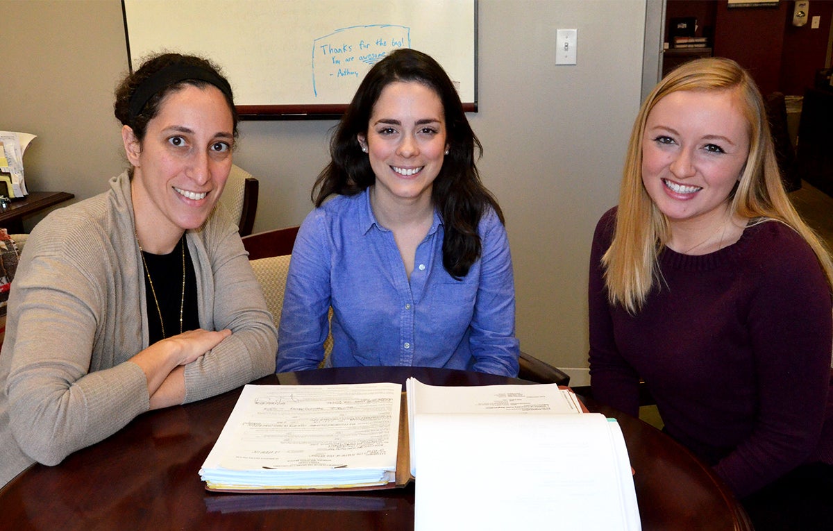 Three women sit side by side at a table, a book open before them.