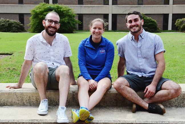 Three students sit on a low wall by a green lawn