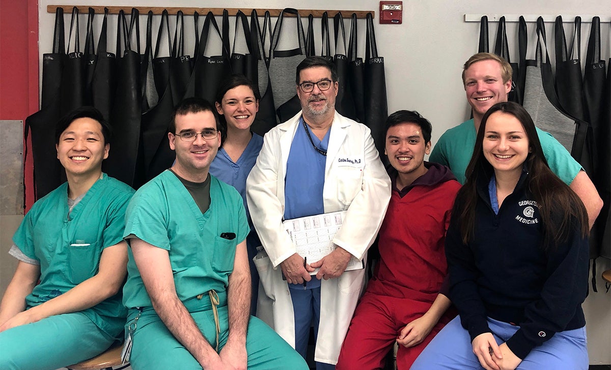 A group of medical students sits with their professor in a group image. Behind them are rows of aprons used in the lab.