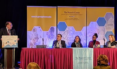 Panelists sit at a table onstage, while a man standing at a podium addresses them
