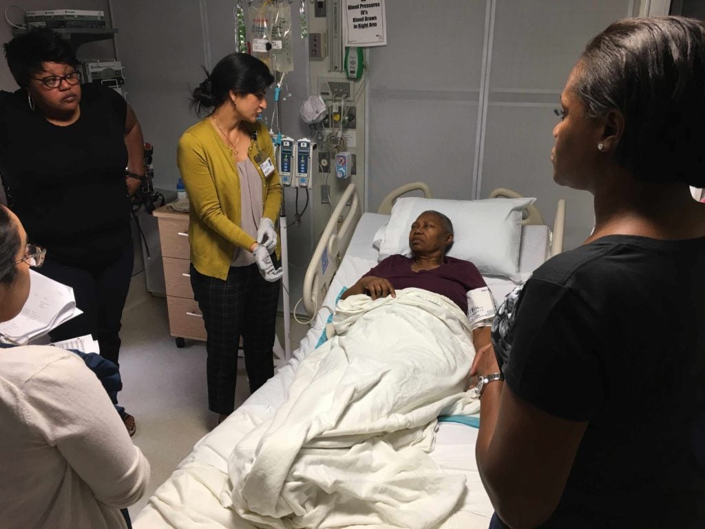 A patient rests in bed while family members and a caregiver stand by her side.
