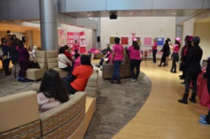 A group of people dressed in pink listen to a woman giving remarks.
