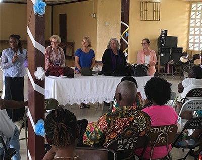 Several women stand behind a table before a crowd seated in a room decorated festively