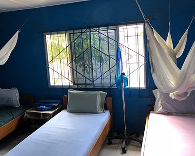 Beds made with pastel-colored sheets are neatly arranged in a room with blue walls and a window. Bed nets hang from the ceiling.