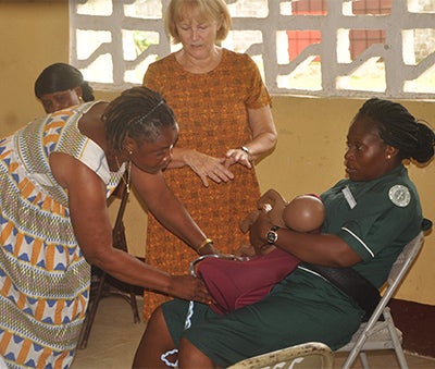 A woman sits with equipment simulating a postpartum woman on her lap, while another woman does a mock exam, guided by a third woman