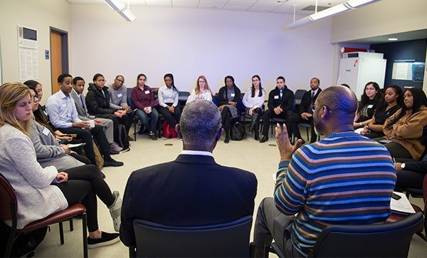 A room ringed with students in chairs looking at two African American men speaking.