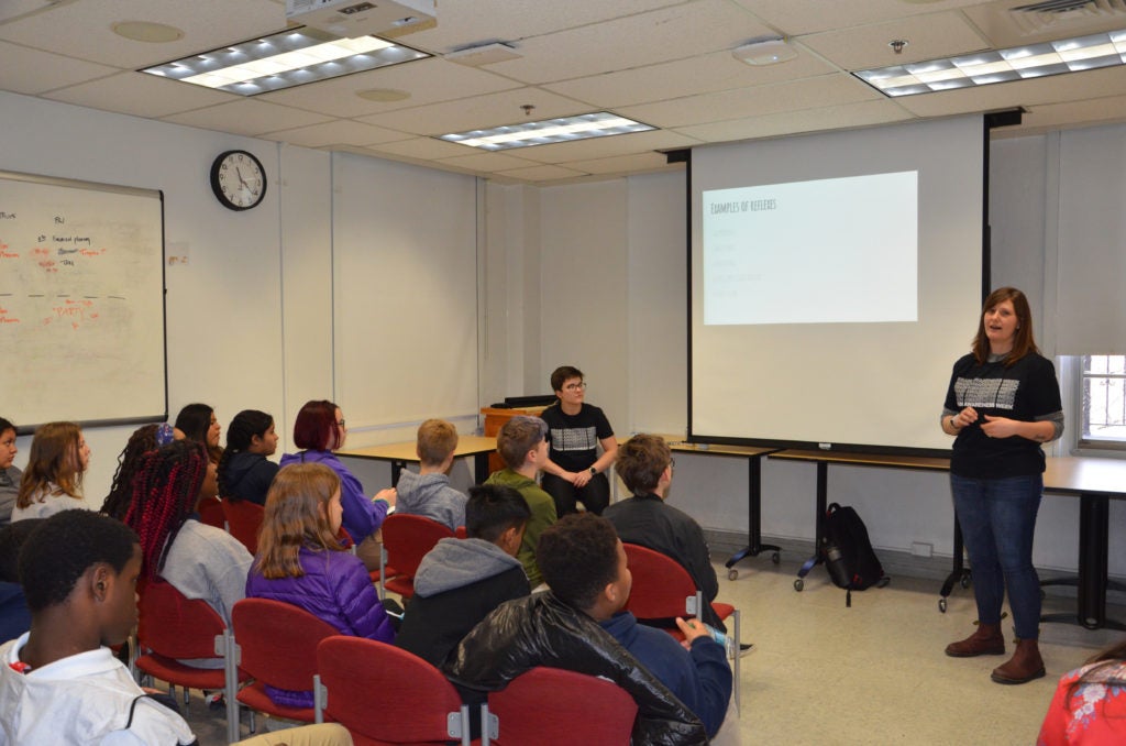 A woman lectures in front of a class full of students.