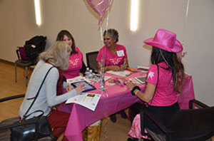 A group of women wearing pink share laughter as they complete paperwork.
