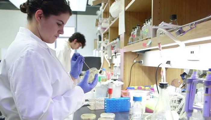 A female student in a white lab coat works in a lab surrounded by equipment