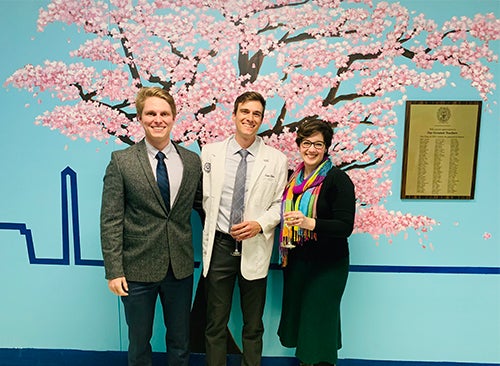 Three individuals stand side by side in front of a mural of a cherry tree in full pink blossom.