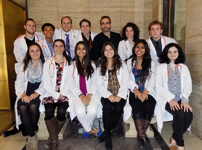 A group of medical students wearing white coats sit in two rows for a group photo