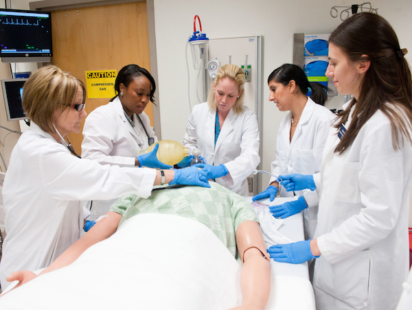 A group of nursing students and their instructor stand around a patient simulation dummy in a hospital bed.