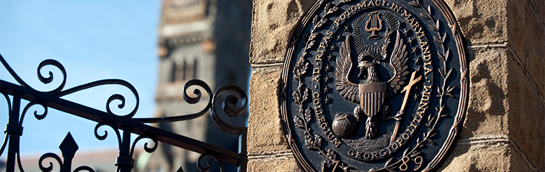 The Georgetown University seal on a brick pillar of a gate with the clocktower of Healey Hall in the background
