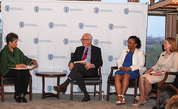 Four individuals sit in chairs before a white drape emblazoned with the GUMC logo