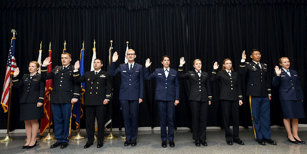 A row of nine people stand in military uniform with right hands raised