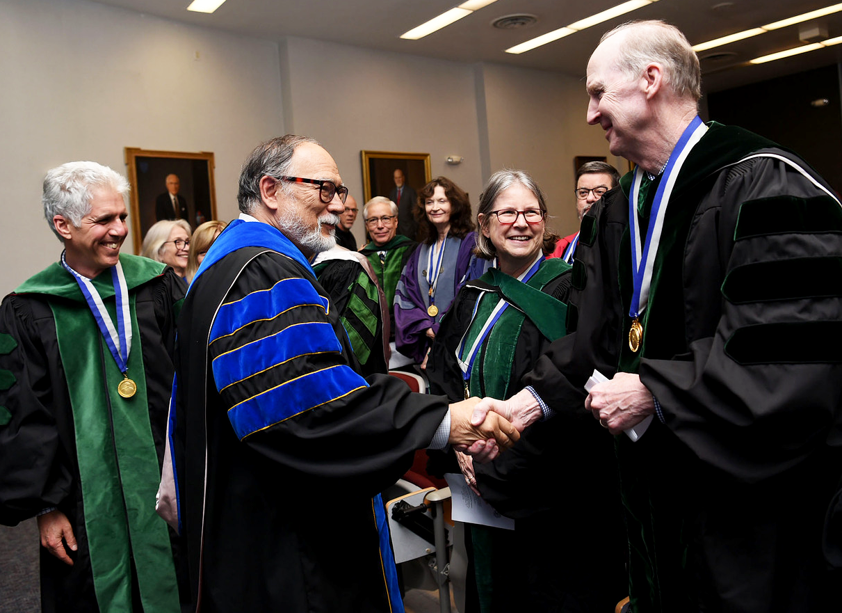 A group of people in academic regalia watch as two men shake hands.