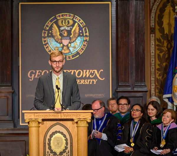 Billy Hoffman speaks at a podium while professors in academic regalia sit behind him