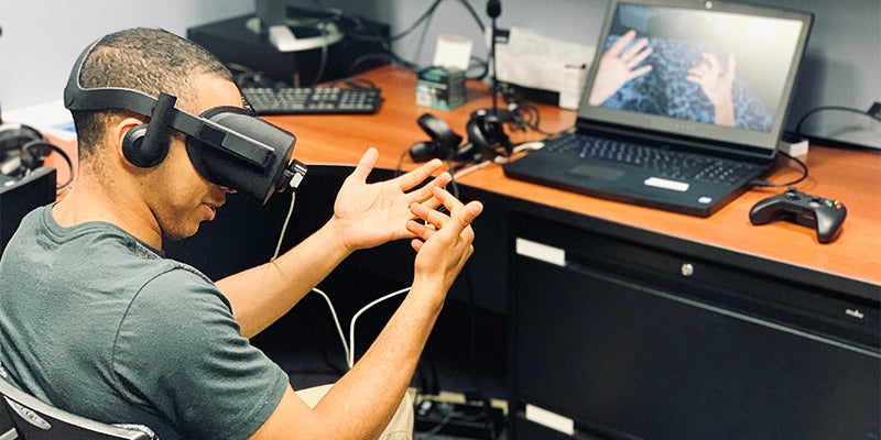 A person wearing virtual reality gear sits in a chair with hands raised before him in front of a computer screen