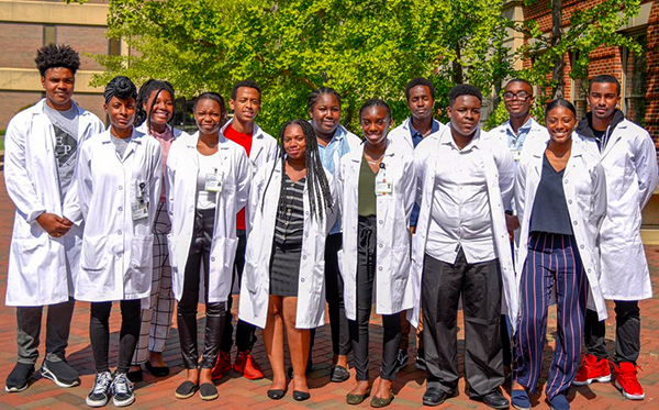 A group of students stand in a row outdoors; all are wearing white lab coats