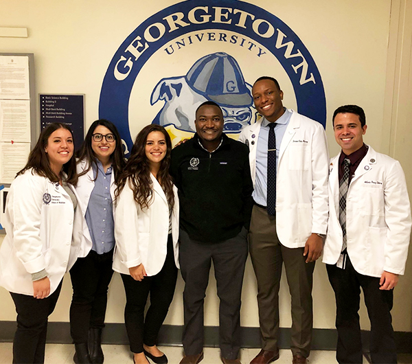 A group of students in white coats with Dr. Kaingo at the center stand before the GUSOM logo painted on a wall in the hallway at the medical school.