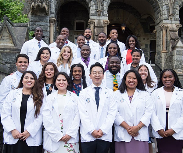 A group of medical students in their white coats stand for a photo on steps at Georgetown