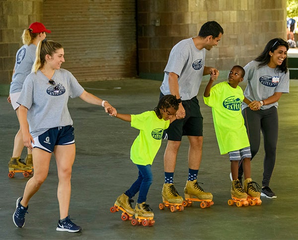 Camp counselors hold the hands of young campers roller-skating tentatively at an outdoor skate park