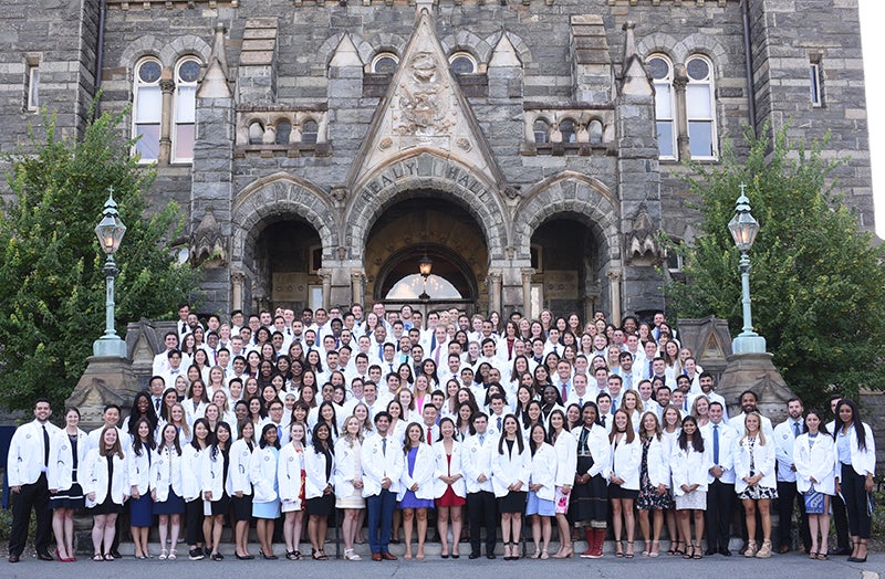 A large group of white coated medical students stands outside Healy Hall