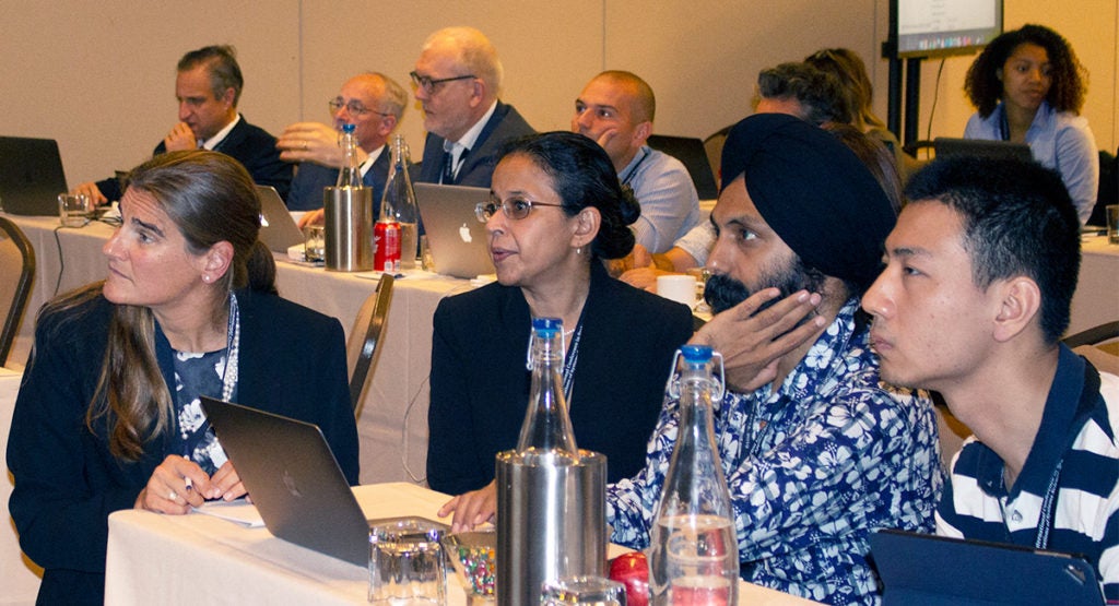 Four individuals sit at a table, with others in the background, looking at a presentation