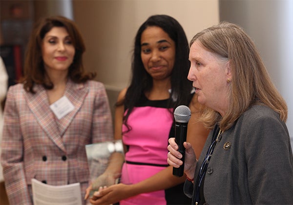 Three women stand together, one holds a microphone and speaks