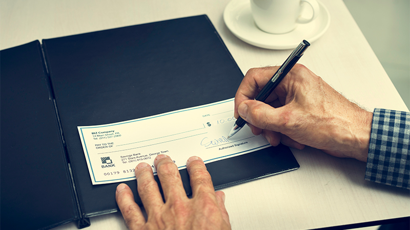 A person's hands are pictured signing a check on a table near a coffee cup