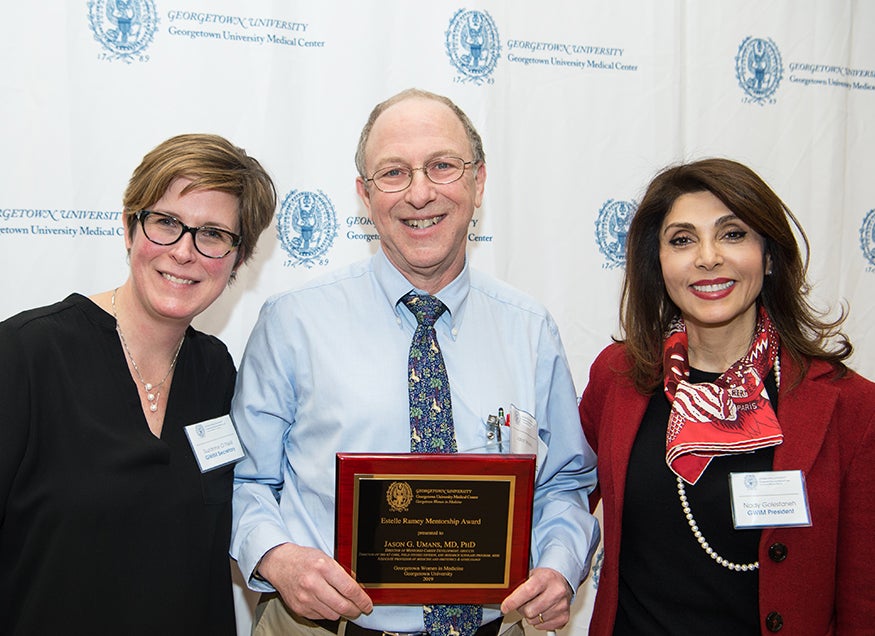 Three people stand together, the person in the middle holds a plaque