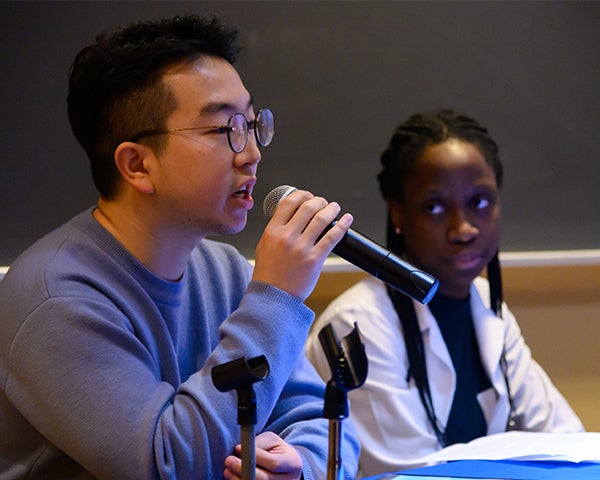 Two people sit at a table, one speaks into a microphone as the other looks on