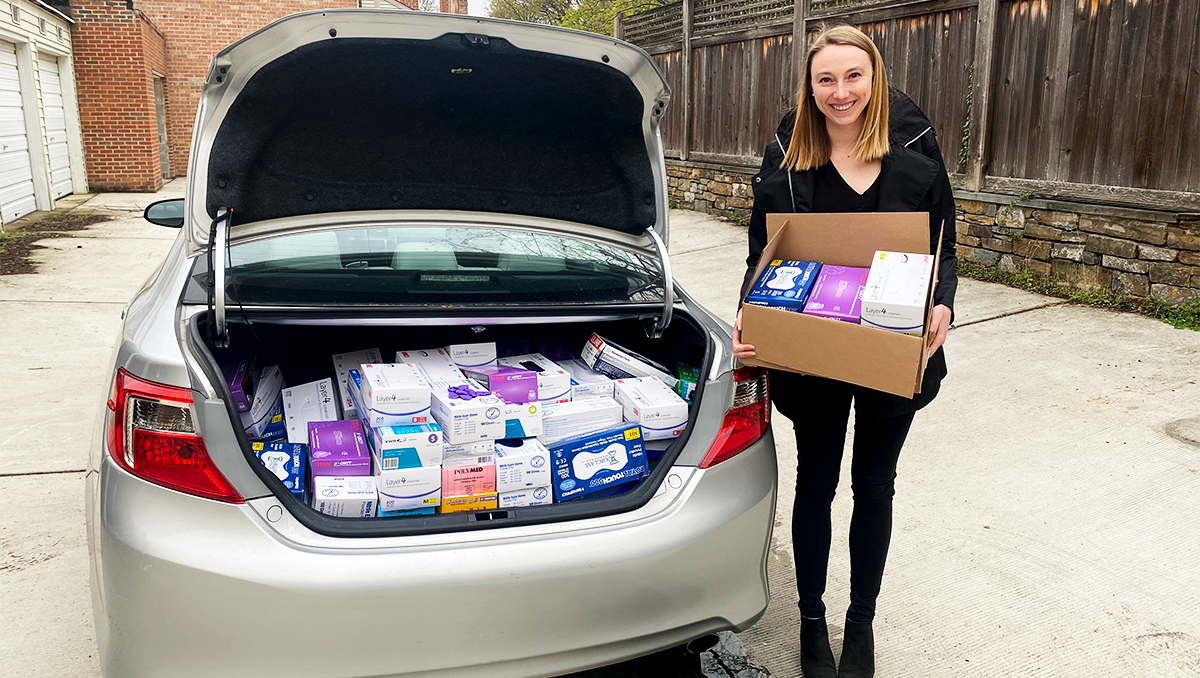 Allison Rooney stands holding a box next to a car with open trunk filled with medical supplies