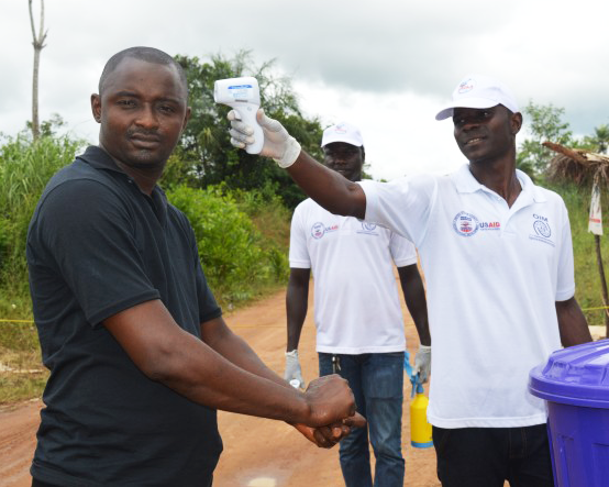 A man at a border crossing in Africa holds a thermometer to the forehead of another man who is washing his hands