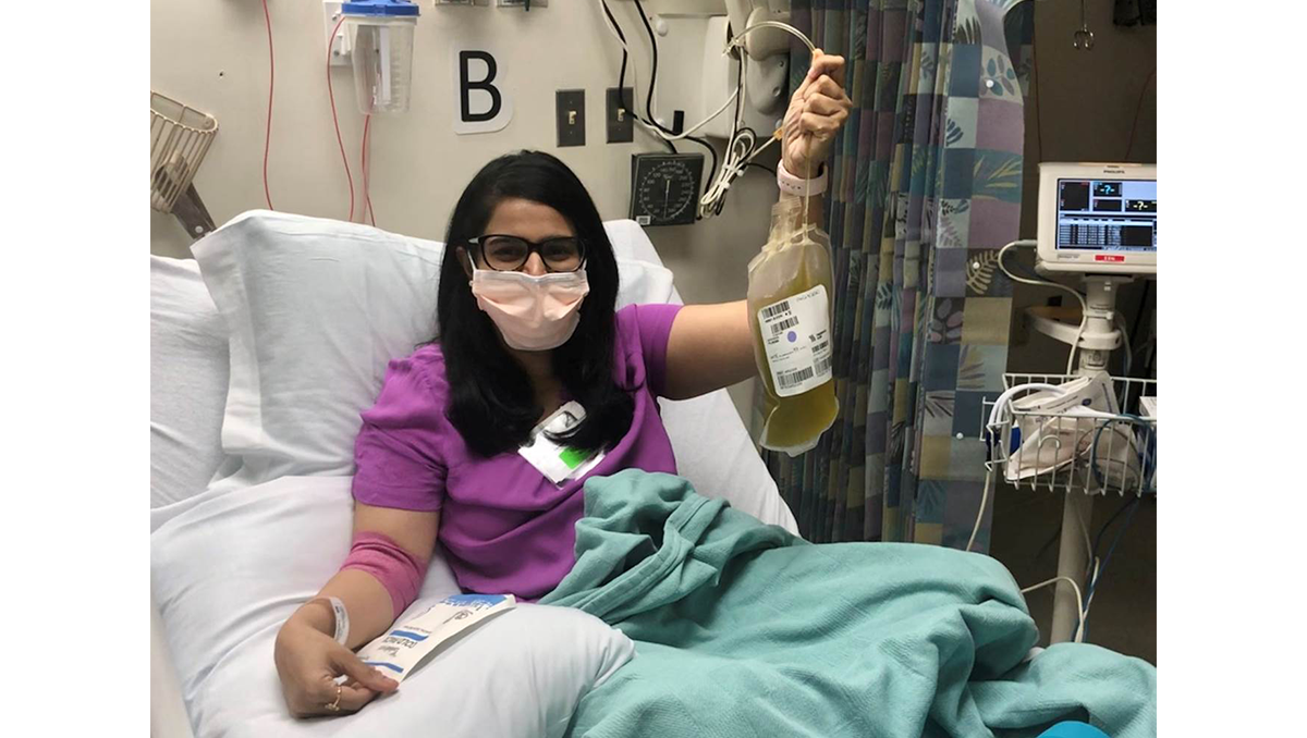 A woman wearing a surgical mask lies in a hospital bed holding a bag of her plasma