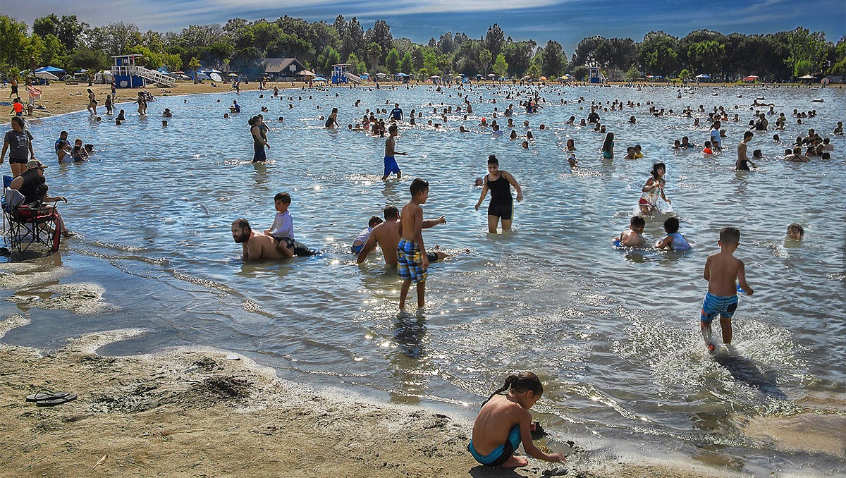 A large number of people swim in a lake and play on the beach without social distancing or masks